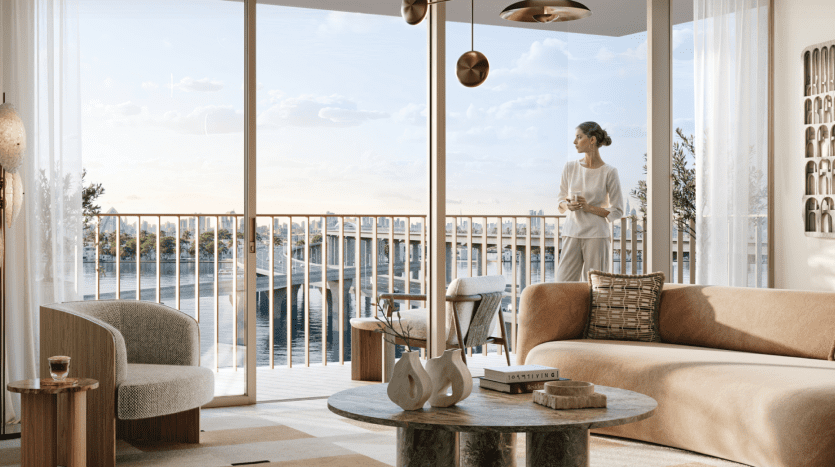 One person stands on a balcony at Address Residences, overlooking a serene waterfront. Inside, the living room features elegant furnishings such as a beige sofa, a green marble coffee table with decoration, an upholstered chair and elegant light fixtures. Large windows let in plenty of natural light.