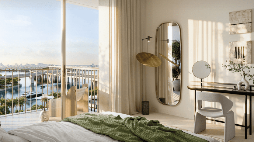 A modern room at Address Residences features a large window offering stunning views of the city's waterfront rooftops. The room features a bed with a green throw, a large oval mirror, a desk with a round mirror and a white chair. Natural light fills the space, creating a serene ambience.