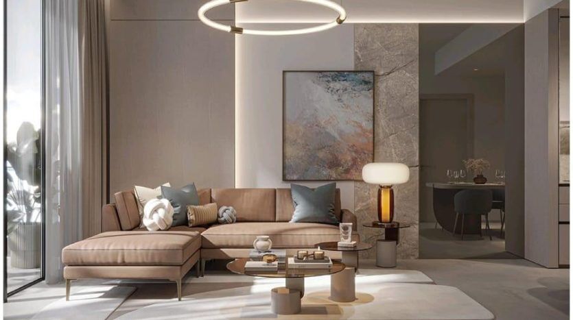 Un salon moderne aux tons neutres comprend un canapé Legado Prescott beige en forme de L orné de coussins, une table basse ronde et un grand tableau abstrait sur un mur d'accent en pierre. L'éclairage comprend un lustre circulaire et une lampe de table contemporaine.