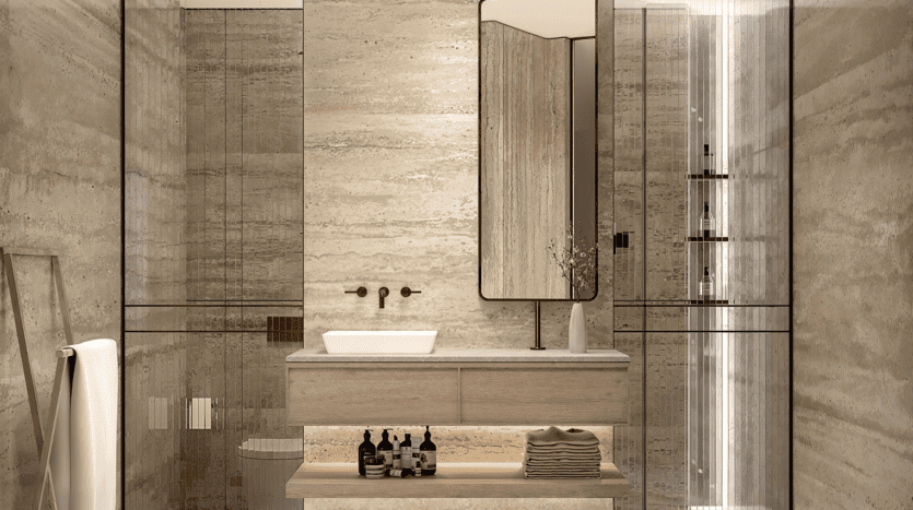 Une salle de bains moderne au One Beverly Dubai avec des murs et un sol en marbre beige. Elle dispose d'un miroir rectangulaire au-dessus d'une coiffeuse en bois clair avec un lavabo blanc. Sous la coiffeuse se trouvent des serviettes pliées et des produits de toilette. À droite se trouve une cabine de douche en verre, tandis qu'à gauche se trouve une échelle porte-serviettes.