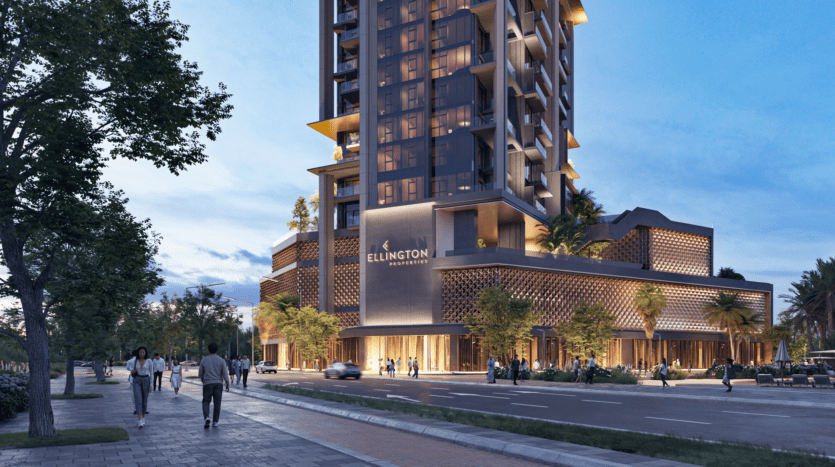 A modern high-rise building with the inscription "Ellington Properties" on the facade stands along a city street. The evening sky is blue and people walk along the sidewalk surrounded by trees and lit by street lamps.