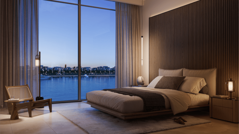 Une chambre moderne du Bay Grove Residences dispose d'une grande fenêtre vitrée présentant l'horizon de la ville au crépuscule. La chambre comprend un lit plateforme avec une literie grise et blanche, une chaise en bois et un éclairage doux. À l'extérieur, les bâtiments se reflètent sur la rivière ou le lac calme.