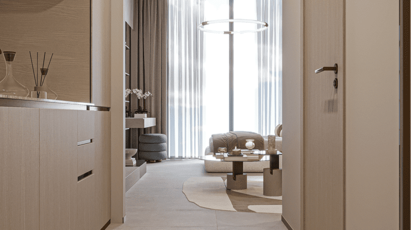 Un couloir intérieur moderne mène à un salon avec une grande fenêtre, des rideaux transparents, un lustre circulaire, des meubles beiges et un tapis à motifs. Des bâtons parfumés et des objets décoratifs sont posés sur une table d'appoint.