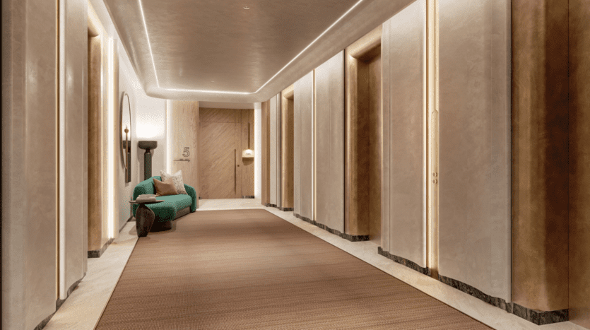 Le couloir moderne du W Residences est doté d'un éclairage tamisé, de murs beiges et d'un tapis beige moelleux. Un canapé bleu sarcelle avec des coussins repose sous un miroir rond à gauche, tandis qu'une porte en bois marquée du numéro 5 est visible au bout. Des panneaux muraux élégants rehaussent la sophistication du couloir.