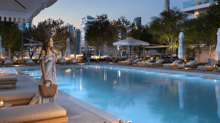 Женщина в длинном платье держит плетеную сумку у бассейна в Marina Cove на закате. В окружении шезлонгов, зонтиков и деревьев на заднем плане виден городской пейзаж, создающий спокойную и роскошную атмосферу.