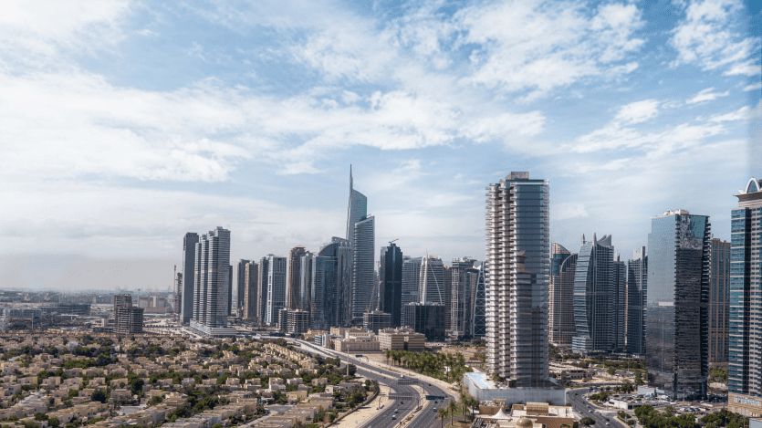 Vue d'une ville moderne avec les emblématiques Jumeirah Lake Towers et de nombreux gratte-ciels de grande hauteur sur un ciel bleu parsemé de nuages. Au premier plan, on aperçoit des routes et des immeubles de faible hauteur, entourés d'une végétation luxuriante.