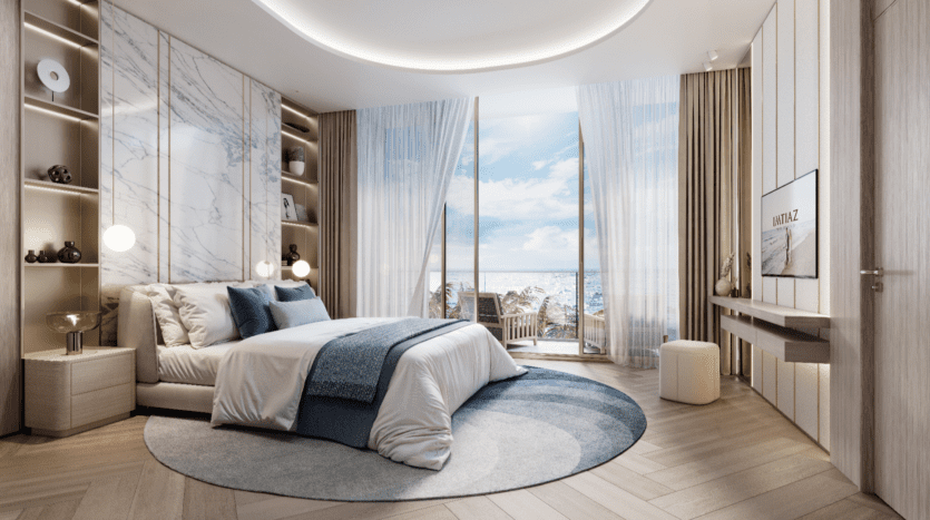 Une chambre luxueuse au Sunset Bay dispose d'un grand lit avec des draps bleus et blancs, d'un tapis rond et de murs aux accents de marbre. Un éclairage doux et des rideaux transparents encadrent une vue sur l'océan à travers une porte vitrée, menant à une terrasse en bois avec des chaises à l'extérieur.