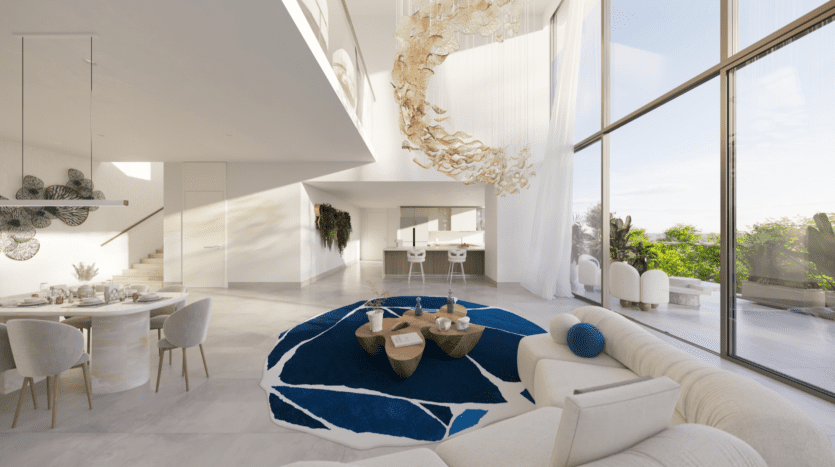 Un salon spacieux et moderne au Rivage avec des baies vitrées laissant entrer la lumière naturelle. Il comprend un tapis bleu et blanc, des meubles beiges, un grand lustre circulaire, un coin repas et une vue sur la verdure extérieure.