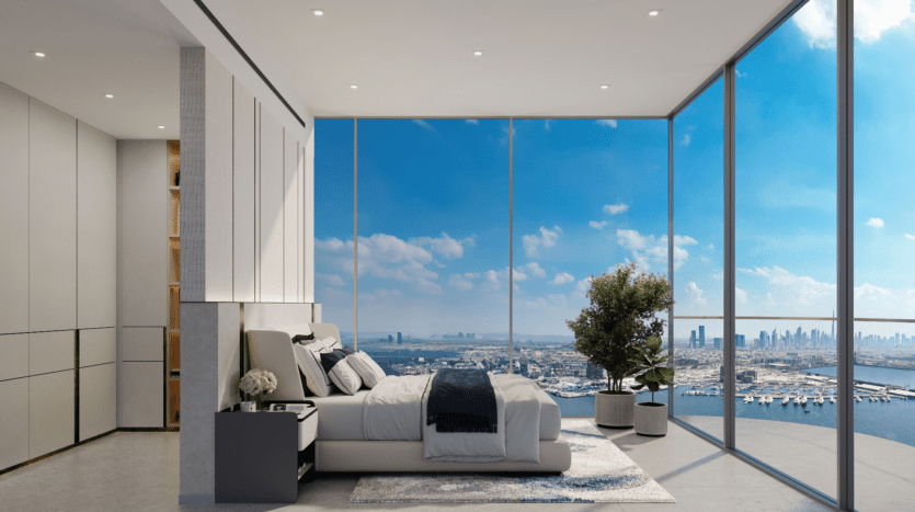 La chambre moderne du LIV Maritime est dotée de baies vitrées qui dévoilent une vue panoramique à couper le souffle sur la ville et l'océan. À l'intérieur, vous trouverez une literie neutre, une table de nuit élégante et une plante en pot aux couleurs vives. Le ciel bleu clair rehausse l'ambiance aérée de la pièce lors de cette journée lumineuse et ensoleillée.
