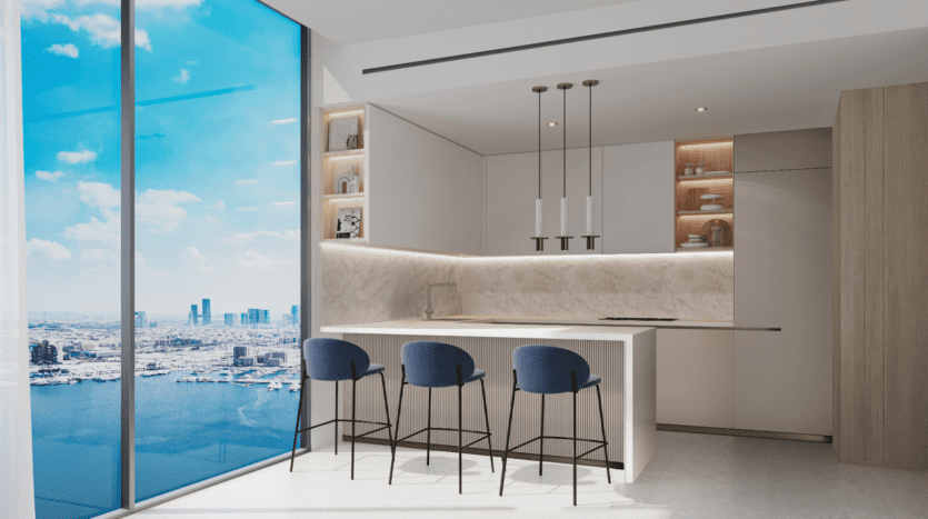 Une cuisine moderne avec des armoires blanches et des comptoirs en marbre comprend trois tabourets de bar bleus sur un îlot, incarnant le style épuré de LIV Maritime. Des luminaires suspendus sont suspendus au-dessus, tandis qu'une grande fenêtre révèle l'horizon de la ville et le ciel bleu avec des nuages en arrière-plan.