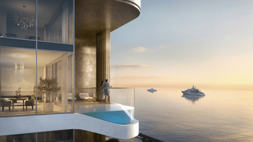 Un couple se tient sur un balcon moderne avec une balustrade en verre au LIV Maritime, surplombant une mer sereine au coucher du soleil. Trois yachts glissent au loin. À travers les grandes fenêtres de l'appartement de luxe, vous pouvez voir une décoration élégante qui complète parfaitement le décor maritime tranquille.