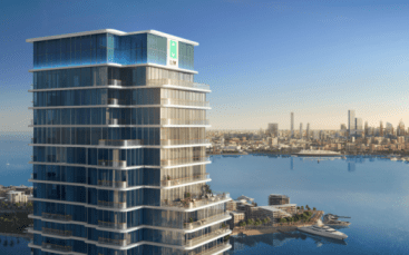 Un immeuble moderne de grande hauteur avec des fenêtres en verre, le LIV Maritime, se dresse en évidence sur le front de mer. La ligne d'horizon de la ville et un célèbre gratte-ciel sont visibles en arrière-plan sous un ciel bleu clair.