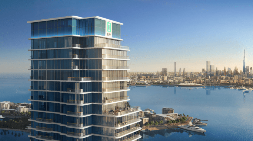Un immeuble moderne de grande hauteur avec des fenêtres en verre, le LIV Maritime, se dresse en évidence sur le front de mer. La ligne d'horizon de la ville et un célèbre gratte-ciel sont visibles en arrière-plan sous un ciel bleu clair.
