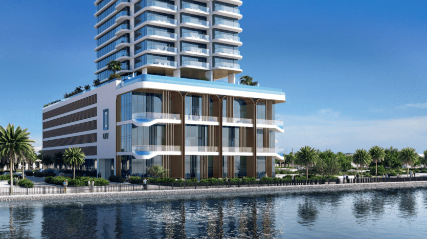 Le LIV Maritime est un bâtiment moderne en front de mer composé de plusieurs étages et de balcons se dressant sur un ciel bleu clair. Sa façade en verre et blanche se reflète dans l'eau calme en contrebas, tandis que des palmiers bordent la passerelle devant cette superbe structure.