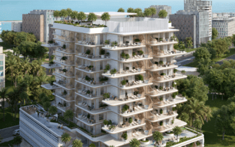 Un immeuble d'appartements moderne à plusieurs étages entouré d'arbres et doté de grands balcons verdoyants se dresse fièrement près de Sunset Bay. La structure offre un aperçu de l'océan en arrière-plan, en harmonie avec les autres bâtiments voisins le long du front de mer tranquille.