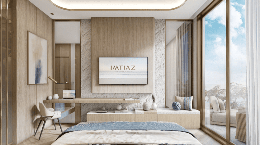 Une chambre d'hôtel moderne de Sunset Bay dispose d'un lit king-size, de finitions en bois clair et d'une grande télévision au mur. Un bureau avec une chaise se trouve sur la gauche, tandis qu'un coin salon confortable près de la fenêtre offre une vue imprenable sur l'océan et le ciel bleu.