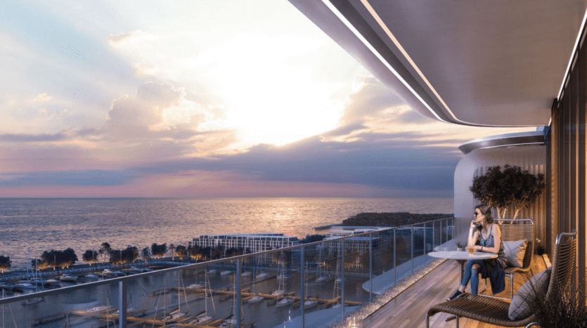 Un balcon moderne de la Cotier House avec une balustrade en verre surplombe une marina au coucher du soleil. Une personne est assise à une table, savourant la vue tandis que des nuages spectaculaires et des rayons de soleil illuminent le ciel et l'océan.