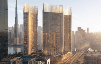 A modern cityscape features three elegant skyscrapers with unique architectural designs, bathed in golden sunlight. These imposing structures overlook a body of water, with roads and smaller buildings surrounding the area.