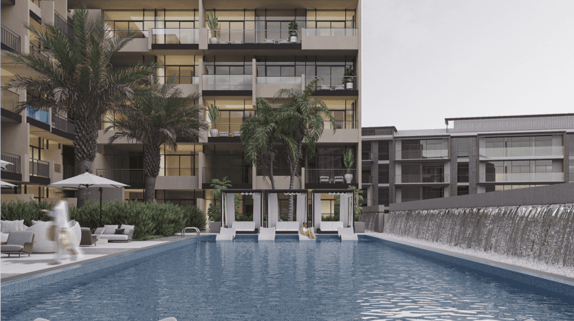 Un complexe d'appartements moderne avec une grande piscine au premier plan, bordée de chaises longues et de palmiers. Une cascade longe le côté droit. Les balcons des appartements donnent sur la piscine.
