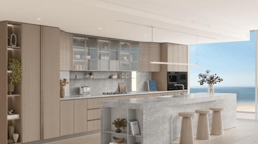 Cuisine moderne à Ellington Cove avec des meubles en bois clair et un grand îlot en pierre. Les armoires supérieures vitrées présentent la vaisselle, tandis que les appareils intégrés et les étagères ouvertes abritent les plantes et la décoration. Vue sur l'océan à travers une fenêtre spacieuse avec trois tabourets en bois près de l'îlot.