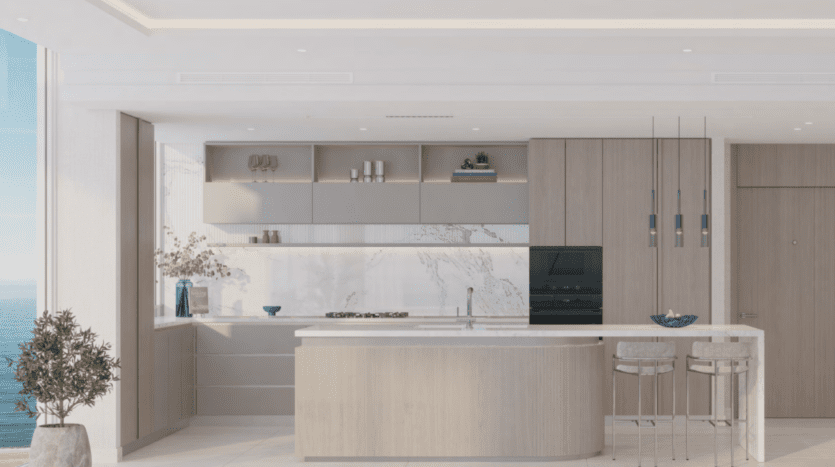 La cuisine moderne d'Ellington Cove est dotée d'armoires en bois clair, d'un dosseret en marbre blanc et d'un îlot avec un comptoir en marbre et deux tabourets de bar. Des étagères intégrées accueillent des objets de décoration. L'espace est lumineux, avec de grandes fenêtres offrant une vue sur l'océan.