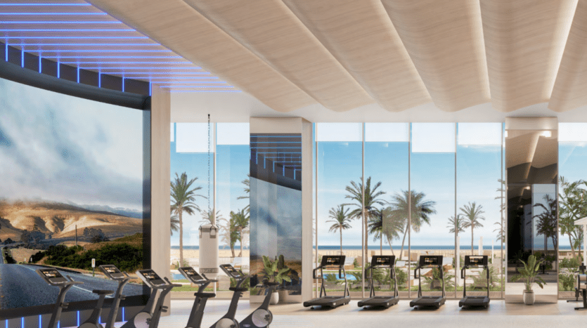 Une salle de sport moderne à Ellington Cove dispose de grandes fenêtres avec des tapis de course parfaitement alignés, offrant une vue imprenable sur les palmiers et la plage au-delà. L'écran incurvé affiche un paysage serein, tandis que les panneaux en bois du plafond et l'éclairage d'appoint créent une atmosphère accueillante.