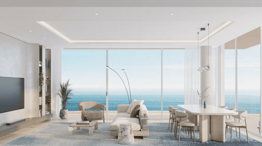 Le salon et la salle à manger modernes de l'Ellington Cove sont dotés de grandes fenêtres offrant une vue sur l'océan. La pièce comprend un canapé de couleur claire, un fauteuil, une table à manger avec des chaises et une décoration minimaliste, comprenant des plantes et un tapis, créant une atmosphère sereine et élégante.