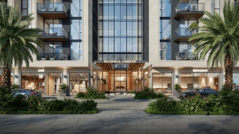 Entrée d'un bâtiment moderne avec de grands panneaux en verre et un panneau « Belgravia Gardens » au-dessus. Flanqué d'un café et d'un restaurant de chaque côté. Des palmiers et une végétation luxuriante bordent l'allée. Deux voitures garées sont visibles près de l'entrée.