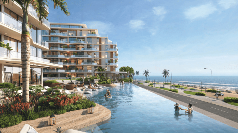 Complexe hôtelier moderne en bord de mer avec une luxueuse piscine à débordement entourée de plantes tropicales. Les clients se détendent dans la piscine et dans les sièges à proximité. Le bâtiment dispose de grands balcons et l'océan est visible sous un ciel bleu clair.