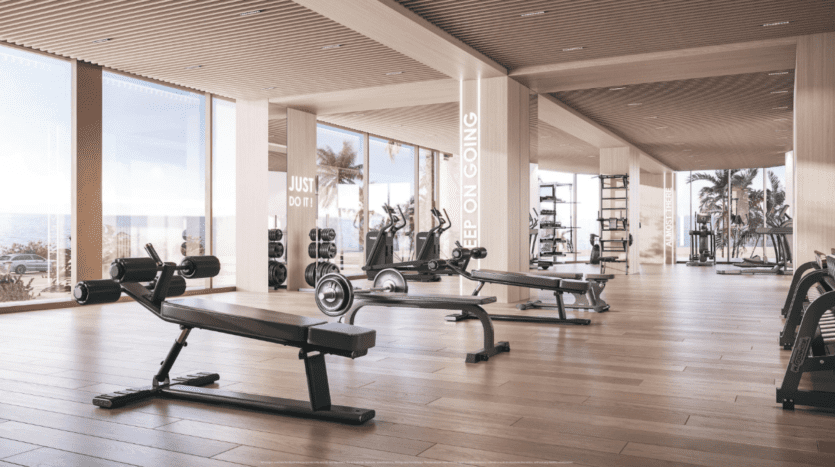 Une salle de sport moderne avec parquet, équipée de bancs de musculation, de vélos d'exercice et de divers équipements de gymnastique. De grandes fenêtres offrent une vue sur les palmiers et l'océan. Des phrases de motivation sont affichées sur les miroirs.