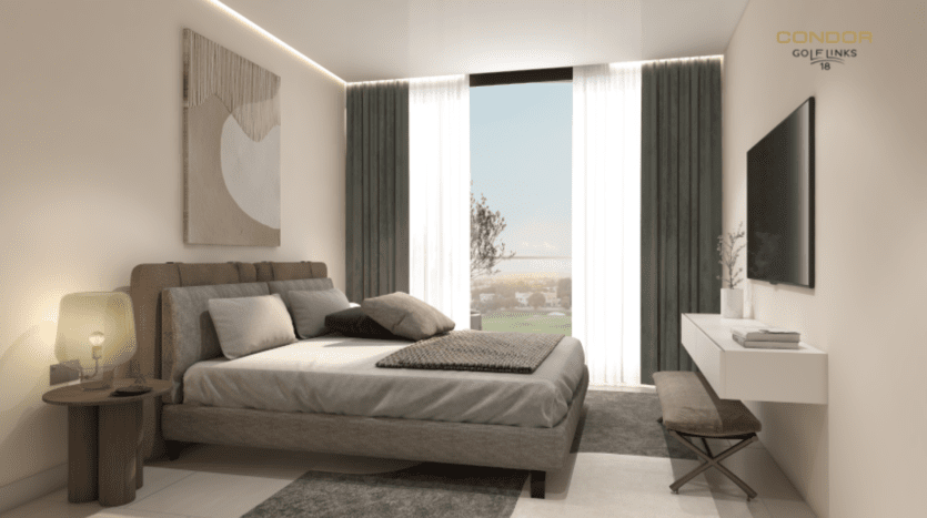 Une chambre moderne dispose d'une grande fenêtre avec des rideaux transparents, permettant à la lumière naturelle d'illuminer l'espace. La chambre comprend un lit avec une literie grise, une télévision murale et un bureau simple idéal pour suivre les parcours de golf. Une tenture murale décorative et une lampe ajoutent des touches esthétiques.