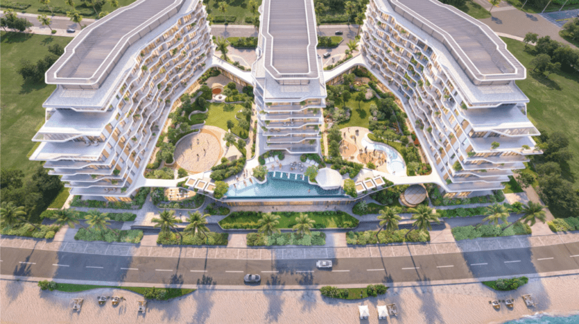 Vue aérienne d'un hôtel moderne de plusieurs étages en bord de mer avec deux bâtiments incurvés. L'hôtel dispose de terrasses, de jardins luxuriants, d'une piscine centrale et de palmiers bordant l'entrée. La plage et une route sont visibles au premier plan.
