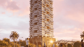 Un gratte-ciel moderne de grande hauteur, au design unique et étagé, se dresse sur un ciel rose et orange au coucher du soleil. Des palmiers et quelques personnes sont visibles au premier plan, ce qui confère une atmosphère animée à la scène.