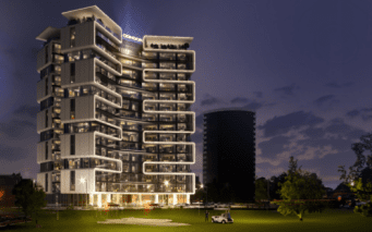 Un bâtiment moderne à plusieurs étages avec des sections éclairées se dresse au crépuscule sur un ciel qui s'assombrit, rappelant la beauté sereine d'un terrain de golf. Les balcons aux formes uniques ajoutent à son charme, tandis qu'au premier plan, une petite voiturette de golf est garée sur une zone herbeuse avec des arbres à proximité.