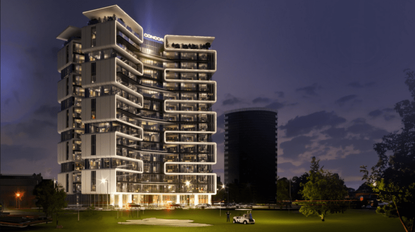 Un bâtiment moderne à plusieurs étages avec des sections éclairées se dresse au crépuscule sur un ciel qui s'assombrit, rappelant la beauté sereine d'un terrain de golf. Les balcons aux formes uniques ajoutent à son charme, tandis qu'au premier plan, une petite voiturette de golf est garée sur une zone herbeuse avec des arbres à proximité.