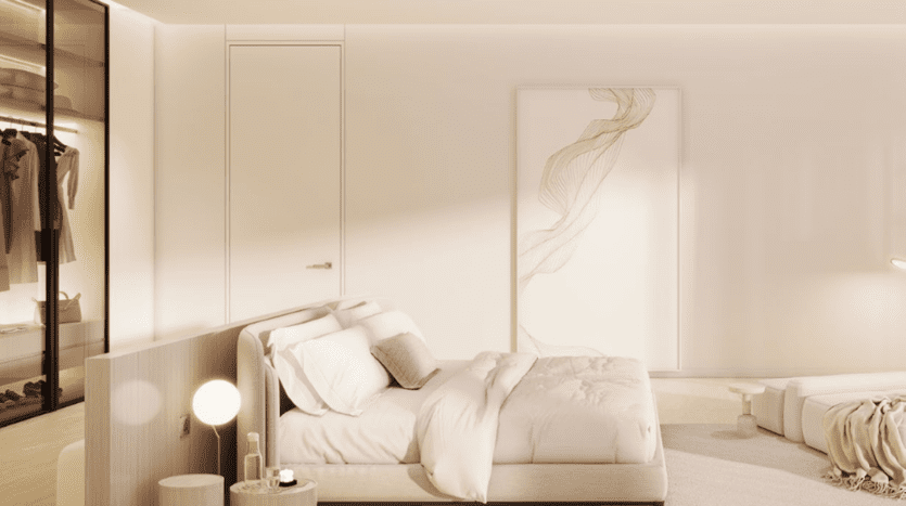 La chambre minimaliste du Chedi Private Residences dispose d'un grand lit avec une literie blanche. Une table de chevet à l'éclairage tamisé accueille une lampe sphérique, tandis qu'une œuvre d'art abstraite aux lignes fluides accentue l'atmosphère sereine. Un placard visible contient des vêtements soigneusement suspendus sur des cintres.