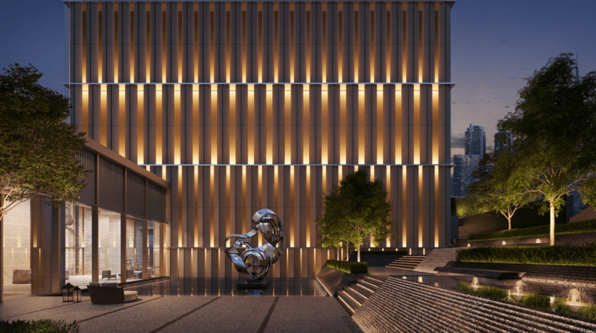 Modern building illuminated vertically at dusk, with reflective abstract sculpture, outdoor space, trees and multi-level landscaping with water features in the foreground. The city's skyscrapers are visible in the background.