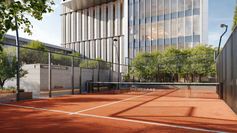 An empty outdoor padel court with a reddish clay surface, surrounded by glass walls and fences, located next to a modern office building with high windows and green trees.
