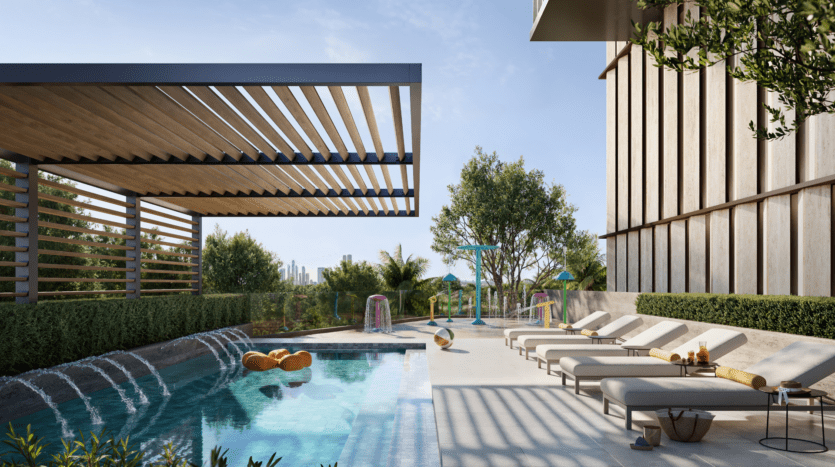 Modern outdoor pool area with water features, sun loungers, shaded pergola, trees and landscaping. Colorful play structures and urban skyline visible in the background under an azure blue sky.
