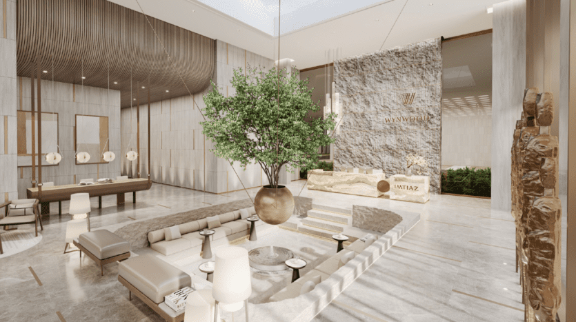 Un hall d'hôtel spacieux et moderne aux tons neutres, avec un grand arbre en pot comme pièce maîtresse, un coin salon en contrebas, un comptoir de réception et des touches de pierre naturelle. Des puits de lumière laissent entrer la lumière naturelle, créant une atmosphère aérée.