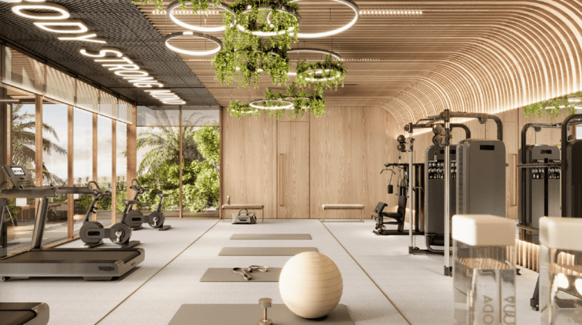 Salle de sport moderne avec murs et plafond en bois clair, grandes fenêtres laissant apparaître de la verdure, équipement d'exercice, tapis de yoga, ballon de stabilité, plantes d'intérieur et luminaires circulaires créant une atmosphère accueillante, lumineuse et aérée.