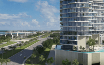 Un immeuble moderne de grande hauteur avec des balcons en verre surplombe une large route bordée de palmiers sur l'île d'Octa, sur l'île de Dubaï, avec des bâtiments plus contemporains et l'océan visibles en arrière-plan sous un ciel partiellement nuageux.