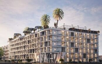 Bâtiment moderne de plusieurs étages doté de grandes fenêtres et de balcons, Avana Residences par Deca Properties à Dubaï se caractérise par des palmiers sur les toits, une végétation luxuriante et des espaces de détente extérieurs, le tout sur fond de ciel bleu vif avec quelques nuages.