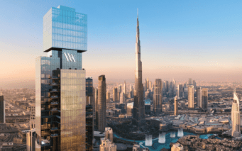 Un grand gratte-ciel moderne portant le logo "Waldorf Astoria" se dresse au premier plan, surplombant un paysage urbain composé du Burj Khalifa et des bâtiments environnants de Dubaï, sous un ciel clair au coucher du soleil.