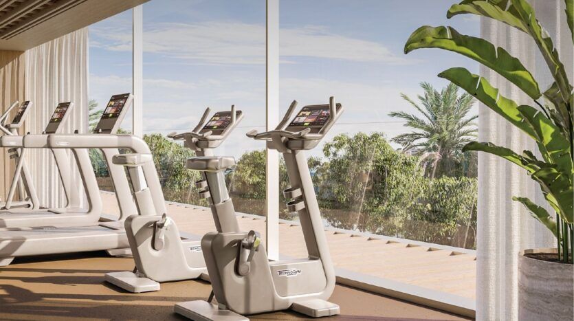 La salle de sport lumineuse de l'Avida Residences Dubai Islands est équipée de vélos d'appartement et de tapis de course qui font face à de grandes fenêtres, offrant une vue panoramique sur des arbres verdoyants et des palmiers. La lumière du soleil remplit la pièce et une grande plante en pot se trouve près de la fenêtre.