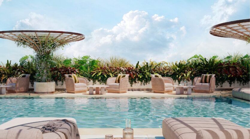 La luxueuse piscine extérieure de l'Avida Residences Dubai Islands, avec des chaises longues, des tables et des fauteuils entourés de plantes tropicales luxuriantes, sous un ciel bleu vif parsemé de nuages.