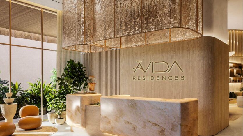 Réception d'hôtel moderne et élégante avec des finitions en bois clair, des comptoirs en marbre, des plantes vertes et de grandes lampes décoratives. Le mur porte l'inscription "ÁVIDA RESIDENCES" en lettres dorées. La lumière naturelle remplit l'espace des îles avida residences dubai.
