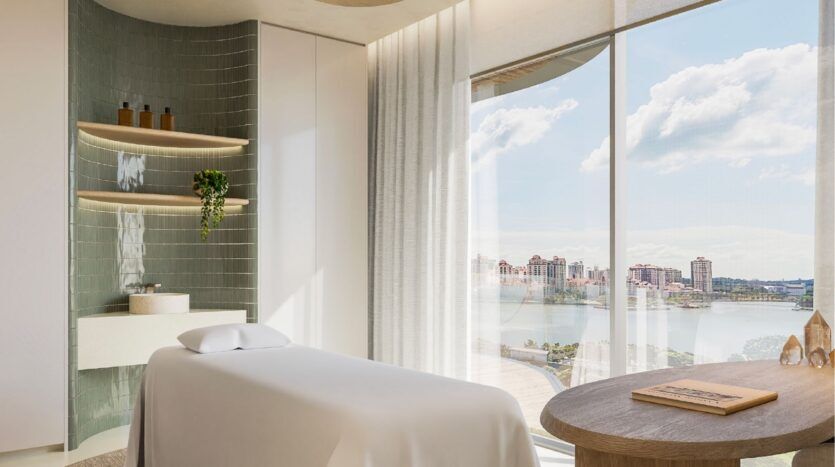 La salle de spa moderne et lumineuse de l'Avida Residences Dubai Islands est équipée d'une table de massage, de grandes fenêtres avec vue sur la ville et l'eau, d'un décor minimaliste, d'étagères avec des bouteilles, d'une petite plante verte et d'une lumière naturelle abondante qui remplit l'espace de détente.