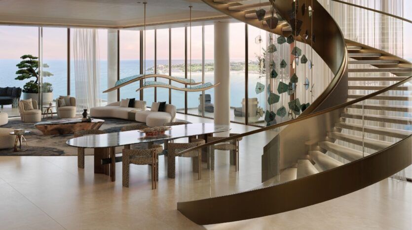 Un espace de vie moderne et spacieux avec des fenêtres allant du sol au plafond et offrant une vue sur l'océan et les îles Silena Dubai, un élégant escalier incurvé, un mobilier contemporain et une grande table à manger, le tout baigné de lumière naturelle.