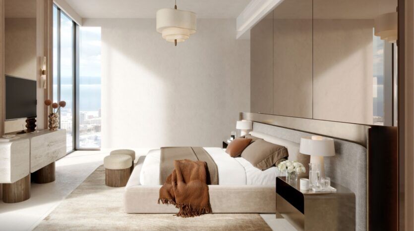 La chambre à coucher moderne aux tons neutres du Portside Square Mina Rashid se compose d'un grand lit avec une couverture brune, de deux tables de chevet avec lampes, d'une télévision sur console, d'une fenêtre allant du sol au plafond et d'une lumière naturelle douce qui remplit l'espace élégant.